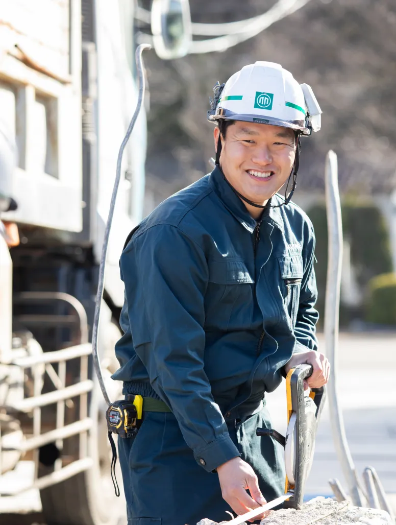 屋外の現場で巻尺を手に持ち、笑顔を見せる青い作業着のT・Kさん