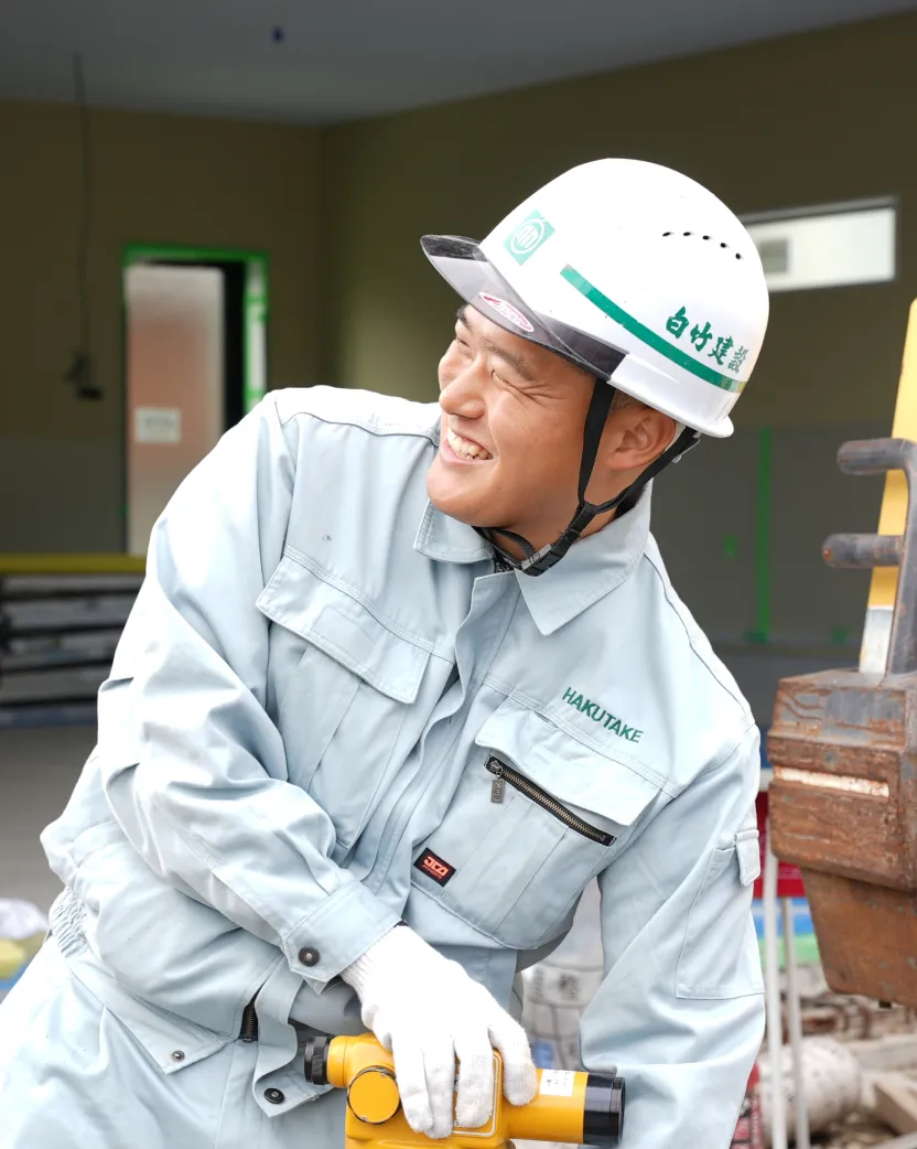 建築現場で黄色い測量機器を手に持ち、笑顔で会話をするヘルメット姿のT・Kさん