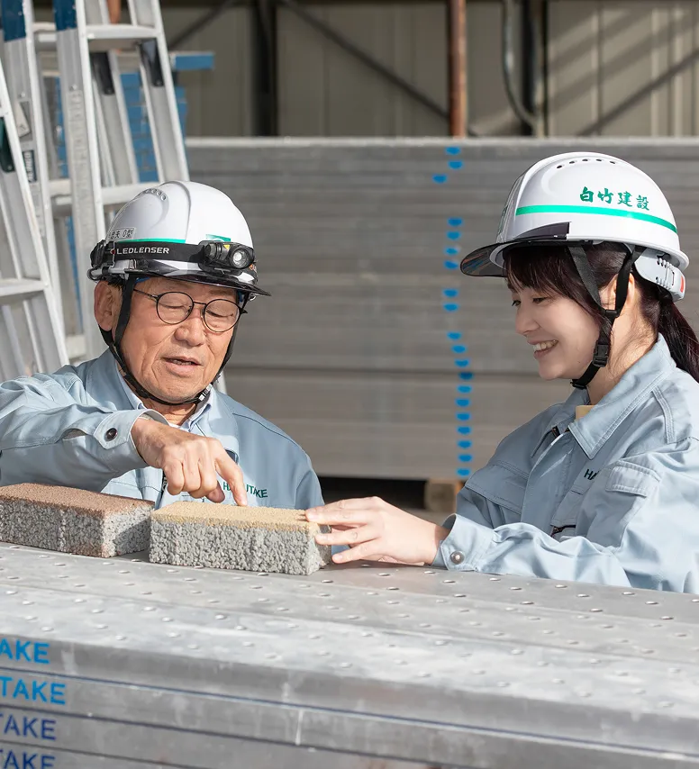若手の女性社員に建材の扱い方について笑顔で指導するベテランのT・Iさん