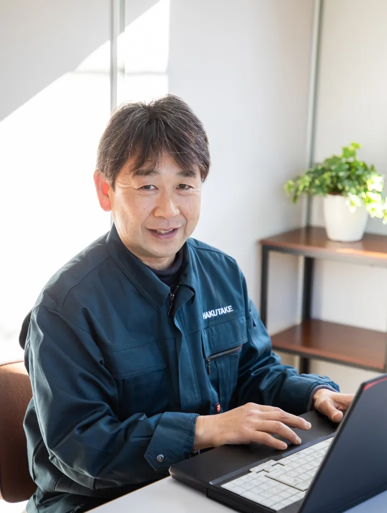 オフィスでノートパソコンを開き、デスクワークを行いながら微笑む青い作業着のT・Sさん