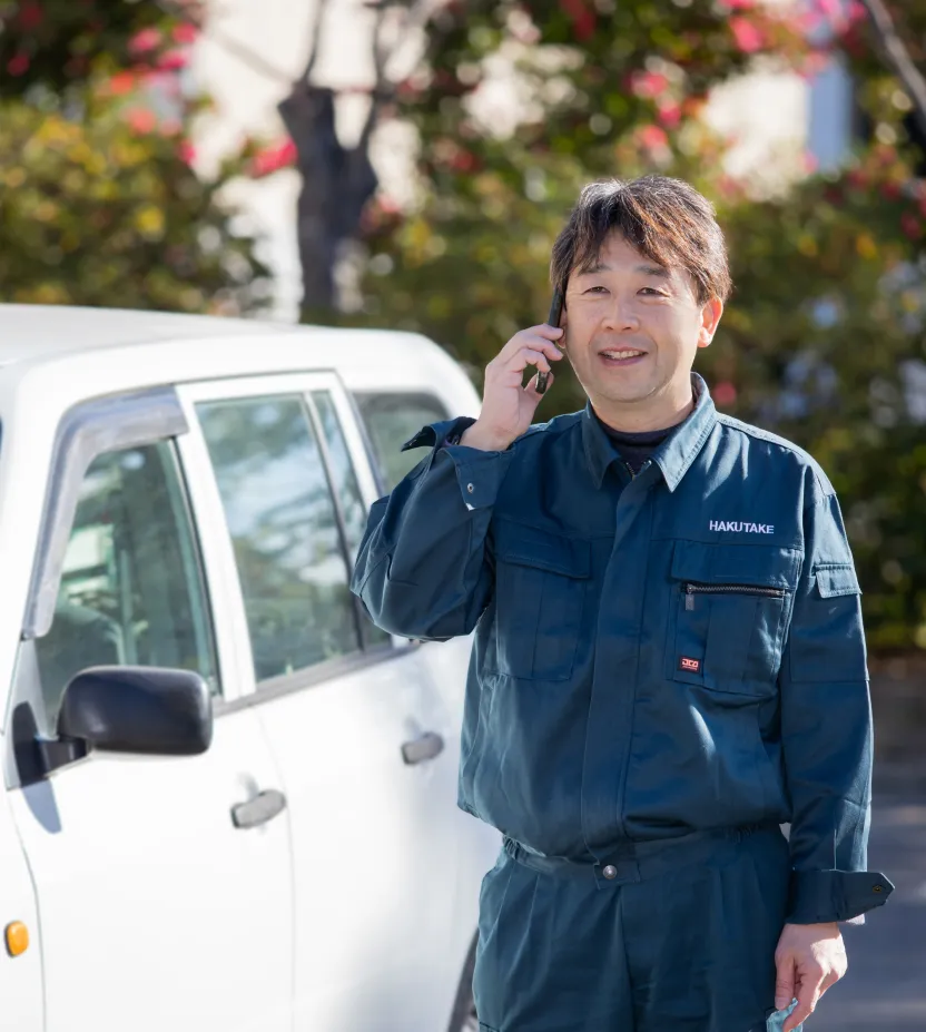 社用車の前でスマートフォンを使い、笑顔で電話連絡をしているT・Sさん
